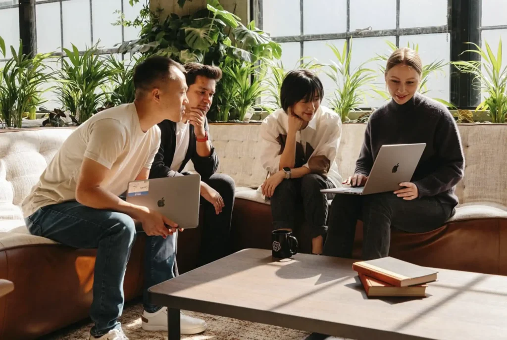 A group of young professionals working on their laptops on a sofa in the common area of Café Hotel Medellín, surrounded by natural light and plants.